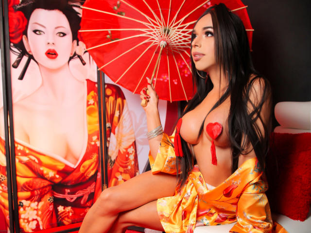 Jhovanna, avec ses longs cheveux noirs, portant un kimono en soie orange, laissant entrevoir ses seins avec des pasties rouges en forme de cœur, tenant un parapluie rouge, assise à côté d'un portrait d'elle-même..