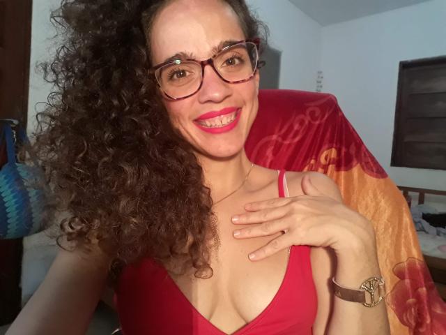 AmyFlores with curly brown hair, red glasses, and red tank top, smiling, showing her tits, hand on chest, pink lipstick, brown wristwatch, and a red towel in background.