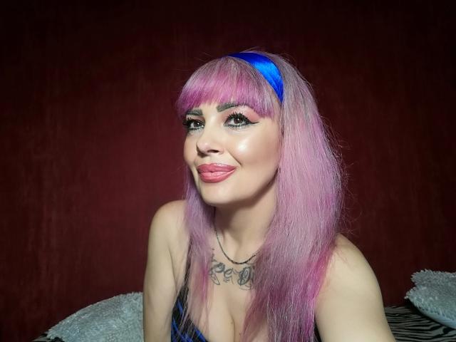 HotAdelayde69 has pink hair with a blue headband, tongue out, and a tattoo above her tits. She's wearing a blue top with black straps.