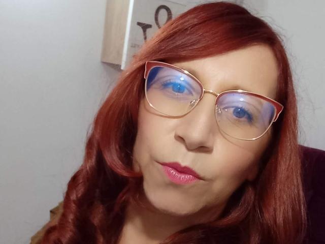 ValeryySweet with red hair, wearing gold glasses, pouty pink lips, and blue reflective eyes, looking slightly goofy. Her tits are not visible, only her face is shown.