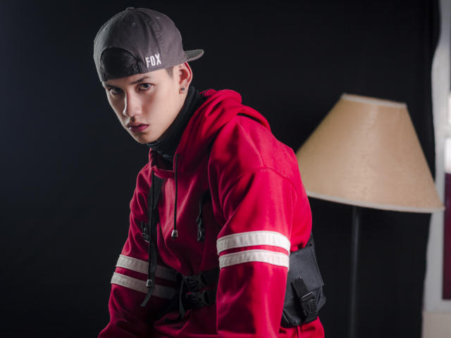 JeanKarloLeone wears a red hoodie with white stripes, black backpack, and gray backwards cap. He has a serious expression, pale skin, and brown eyes. Lamp in the background.