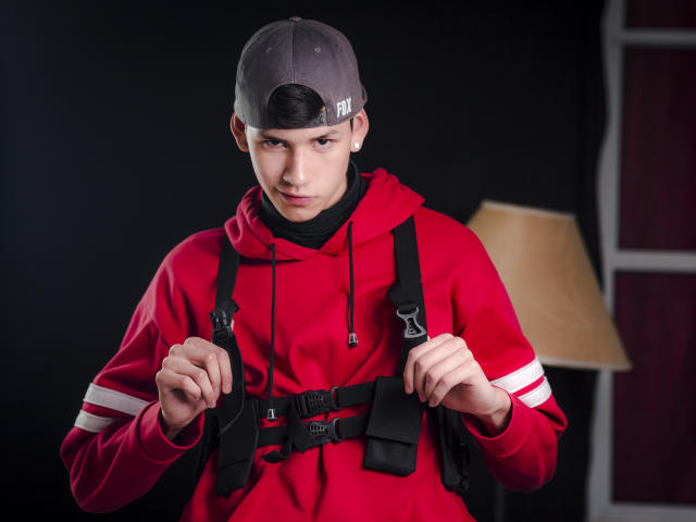 JeanKarloLeone, wearing a gray baseball cap and red hoodie, grips black straps of a tactical vest, with a serious expression, in a studio setting with a lamp.