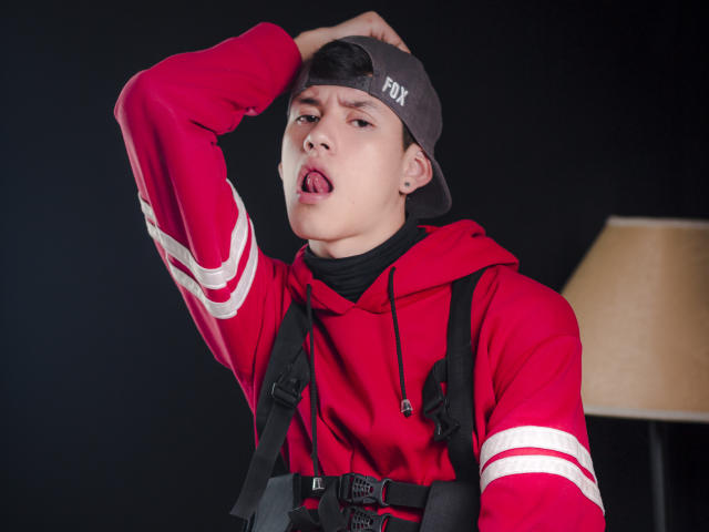 JeanKarloLeone with a cocky expression, tongue out, wearing a red hoodie with white stripes, black backpack straps, and a gray Fox cap, raising his right arm.