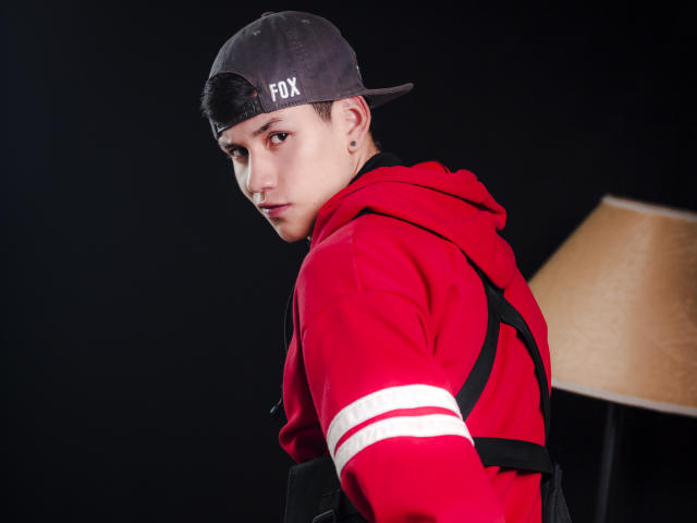 JeanKarloLeone, wearing a red hoodie with white stripes, black straps, and a gray "FOX" cap, looking over shoulder, dark background, lamp on right.