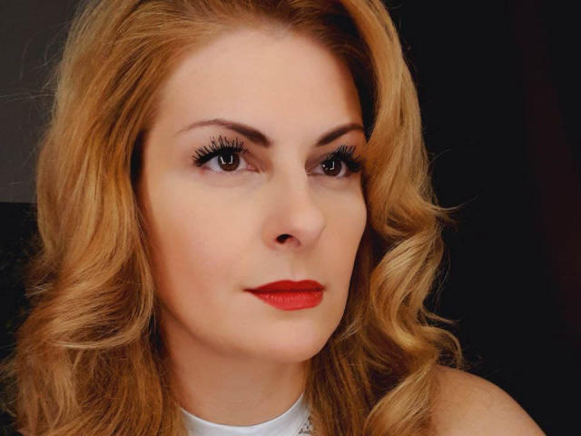 Eleonoress has blonde, wavy hair, red lipstick, and brown eyes with long, dark eyelashes. Her fair skin is smooth, and she's wearing a white top.