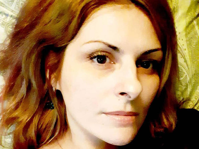 Eleonoress has pale skin, red wavy hair, small nose, brown eyes, and a slight smile. She's wearing black earbuds with a subtle, confident look.