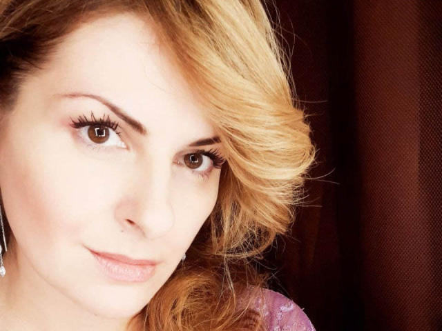 Eleonoress has blonde, wavy hair, brown eyes, and a slight smile. Her pink lips are soft, and she's wearing a purple, shiny top with a single earring.