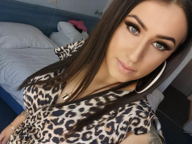 LovelyMichellex69 with long brown hair, green eyes, and a leopard print top, wearing large hoop earrings and a gold necklace, has a tattoo on her right arm.