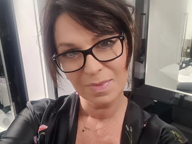 KellyMatureX has short brown hair, black glasses, and is wearing a black floral robe. Her pink lips and subtle makeup are noticeable. She has a gold necklace.