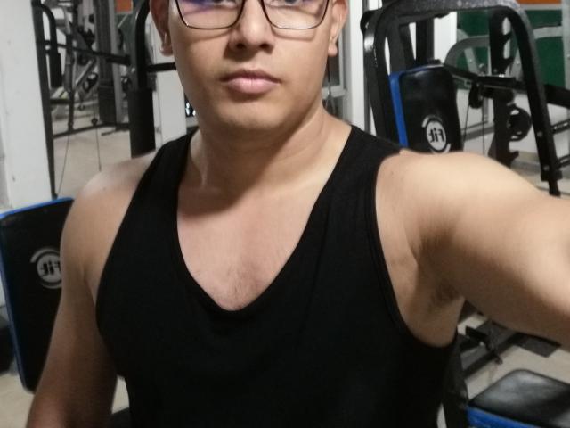 PatrickJean with glasses, black tank top, muscular arms, light brown skin, short dark hair, slightly puffy lips, in a gym, holding a selfie.