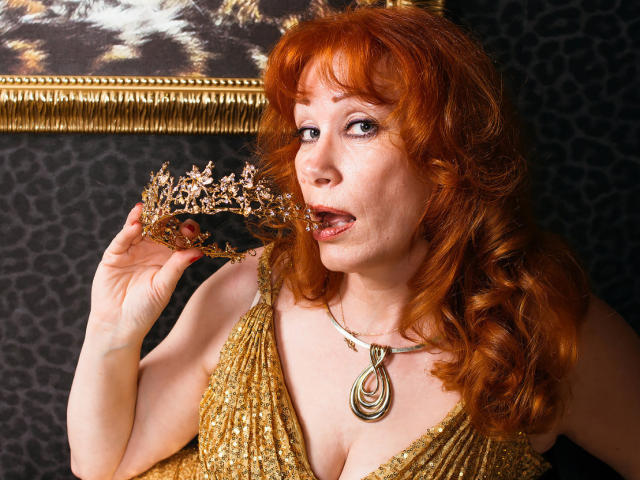 HornyLadiy with red curly hair, gold sequined dress, and necklace, licking a gold and pink floral crown, showing her tits, in a sexy, sultry pose, with a playful expression.
