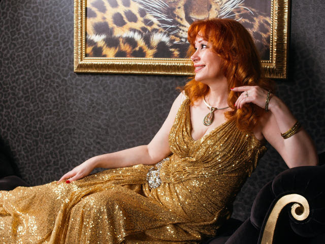 HornyLadiy with red hair, sparkling gold dress, and necklace, sitting on black velvet couch, looking to the side, smiling, hand on chin, showing her ass slightly.