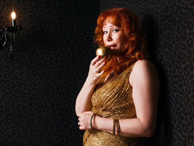 HornyLadiy with red hair, holding a gold apple, wearing a glittery gold dress, standing against a dark leopard print wall, candle in the background, looking playful.