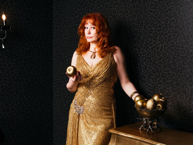 HornyLadiy with red curly hair, wearing a shiny gold dress, holding a golden apple in one hand, and a bowl of golden apples in the other, standing near a wooden table.