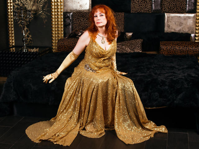HornyLadiy with red hair, wearing a sparkly gold dress, sitting on a black bed, in yellow gloves, showing off her tits and ass, with a sexy, confident look.