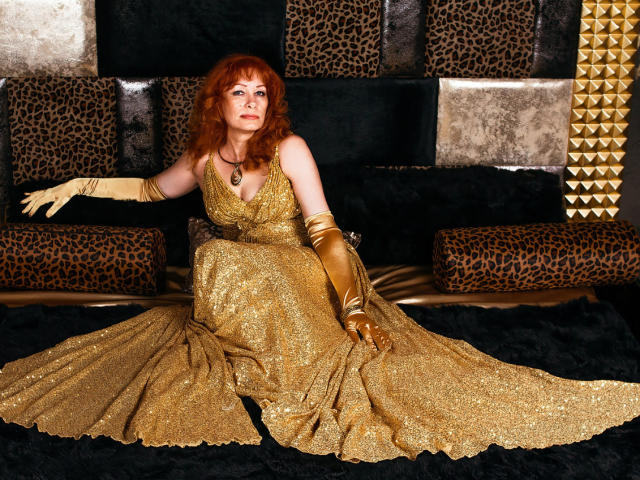 HornyLadiy with red curly hair, wearing a glittery gold dress and gold gloves, sitting on a black fur rug, spreading her legs, showing her ass, and looking up.