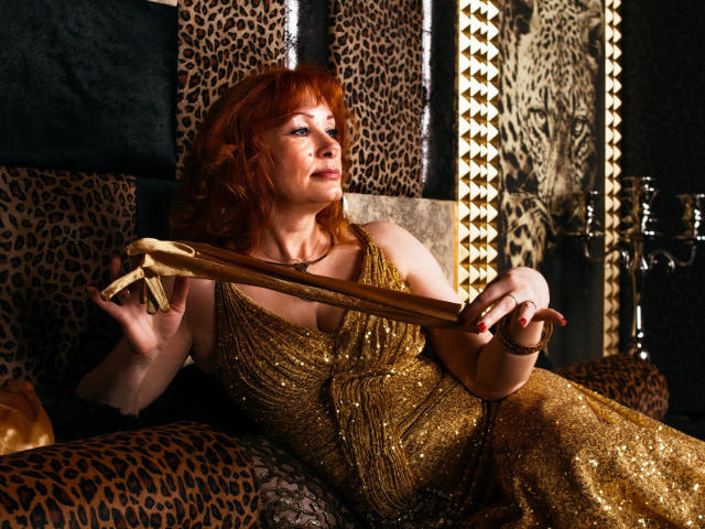 HornyLadiy with red hair, wearing a sparkly gold dress, holding a gold ribbon, sitting on a leopard print chair, looking up with a slight smile.