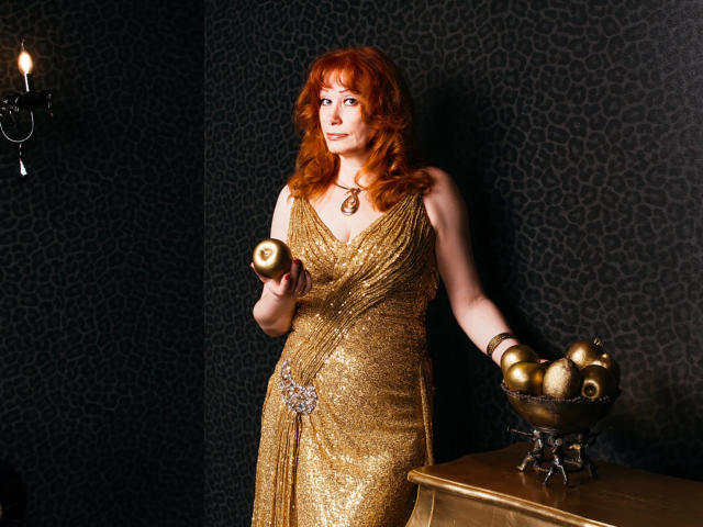 HornyLadiy with red hair, wearing a gold, sparkly dress, holding a candle, standing by a table with a brass candle holder, her tits slightly visible, looking to the side.