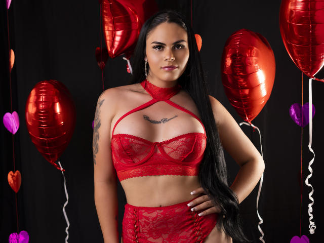 JassmineTS with long black hair, wearing red lace lingerie, has large tits, a tattoo on her right arm, and red balloons in the background. She stands confidently, hand on hip.