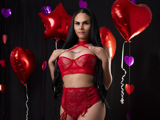 JassmineTS with long black hair, wearing red lace lingerie, holding her tits, standing in front of red and purple heart balloons, looking at the camera with a slight smile.