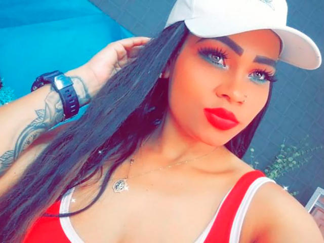 LinaaPrincess with long black hair, wearing a white cap, red and white top, blue eye makeup, red lipstick, wrist tattoo, and black watch, looking at camera.
