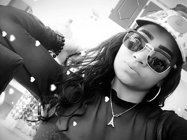 LinaaPrincess with long dark hair, wearing a cap, sunglasses, hoop earrings, and a necklace, has a pouty face and a turtleneck, with her arm raised in a selfie.