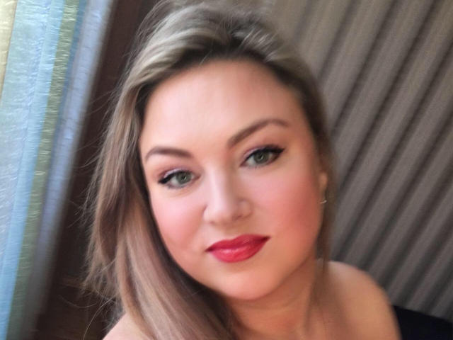 HugeNaturalTits has blonde hair, green eyes, and red lipstick. She's smiling with subtle makeup, wearing a black top. Her tits are not visible. She's in a soft light.