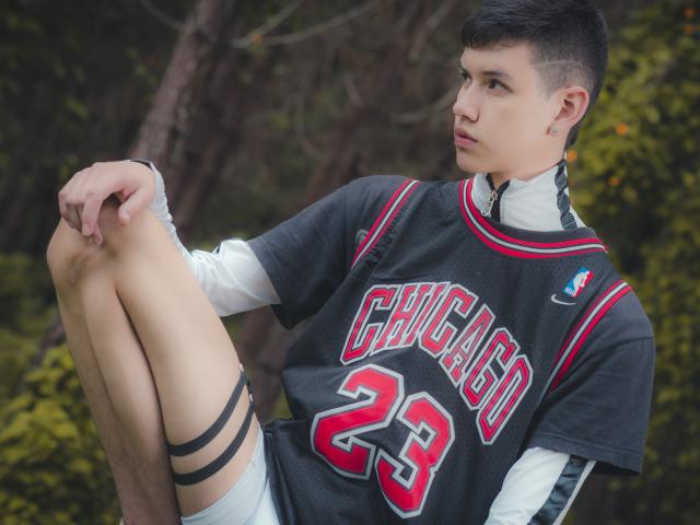 JeanKarloLeone with short black hair, wearing a Chicago 23 jersey, white long-sleeve shirt, and gray shorts, sitting with knees up, looking to the side, in a forest.