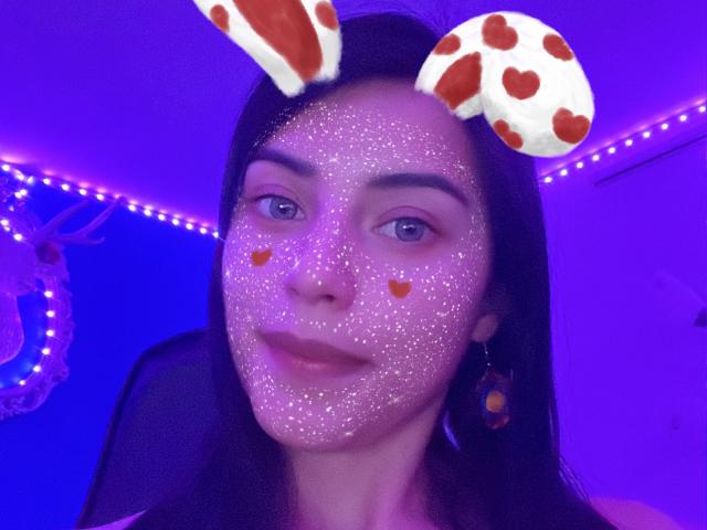 Arenne with glitter on her face, cow ears emoji, orange heart emojis on cheeks, purple LED lighting, black hair, blue eyes, and purple-tinted skin in a selfie.