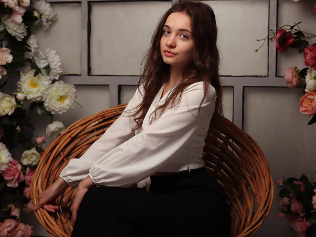 LuiMartin with long brown hair, white blouse, and black pants, sitting on a wicker chair, surrounded by flowers, looking to the side with a subtle smile.