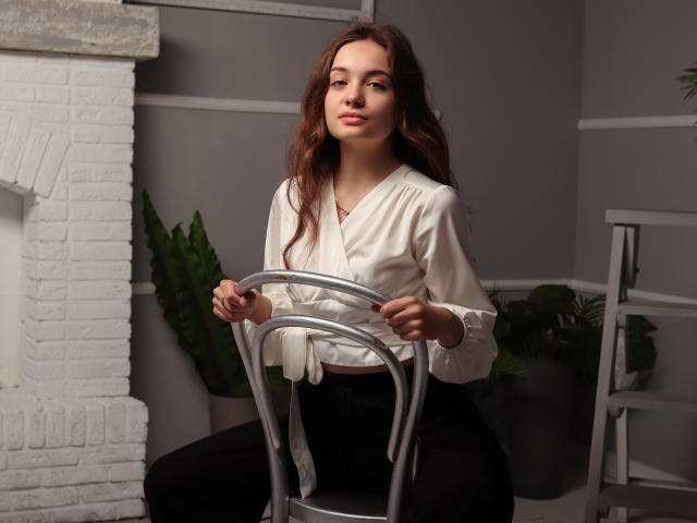 LuiMartin, with wavy brown hair and white blouse, sits on a silver chair, holding the backrest, wearing black pants, in a gray room with white brick and plants.