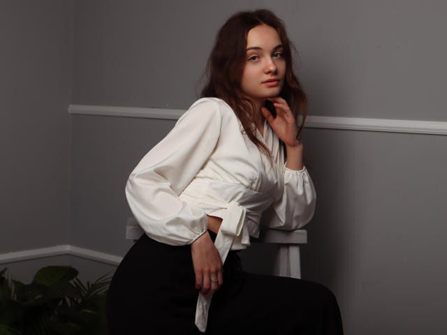 LuiMartin has wavy brown hair, wearing a white blouse with long sleeves and black pants, sitting with one hand on her chin, looking slightly to the side.