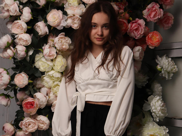 LuiMartin with long brown hair, wearing a white, long-sleeve, tied-up top, black pants, standing in front of a floral wall with pink, white, and red roses.