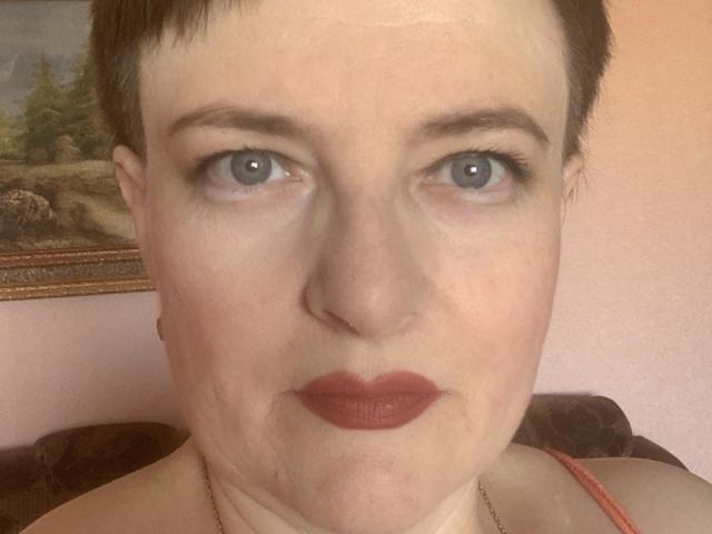 MargieEvans has short brown hair, blue eyes, and red lipstick. Her face shows a slight frown. She's wearing a red strap and a gold necklace.