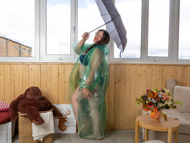 BerryChic with green translucent robe, holding black umbrella, smiling, showing ass, in room with wooden panel walls, red teddy bear on basket, colorful flowers on wooden table.