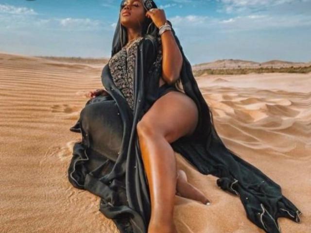 KatyBomb with dark skin, long black hair, wearing a black, patterned dress, sitting on sandy desert, showing her ass, legs crossed, holding black phone, blue sky above.