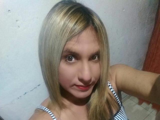JaneStonesTs has blonde hair, light skin, and is wearing a striped tank top, with a slight smile and red lips, in a selfie. Her tits are small and cute.