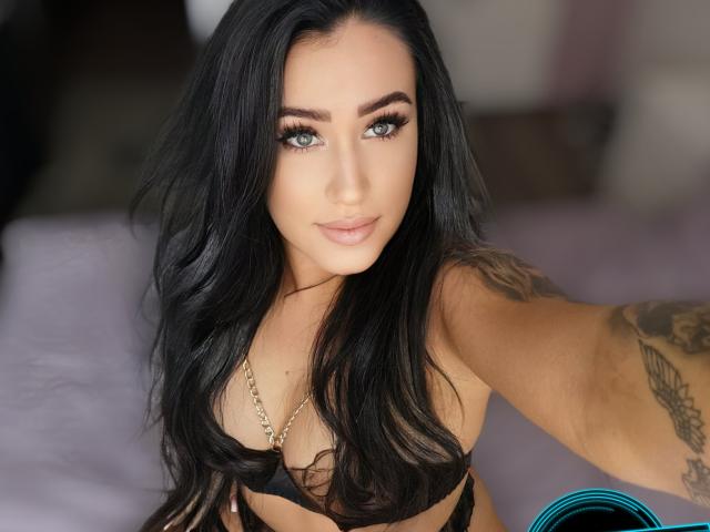 LovelyMichellex69 with long black hair, blue eyes, and tattoos, wearing black lingerie, looking at the camera with a slight smile, showing her tits.