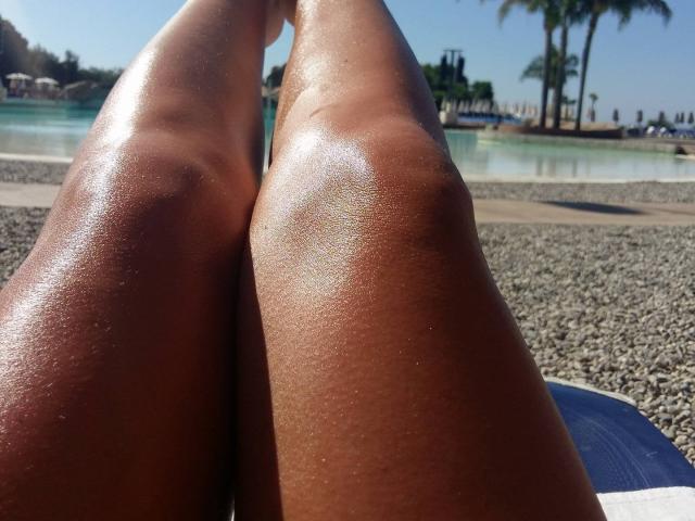 Shiny, sun-kissed, brown legs of JustFuckMeXX, with slight glistening from sweat, spread apart on a pebble-covered beach, showing their smooth, toned thighs in a relaxed position.
