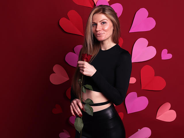 GretyArdent with long brown hair, holding a red rose, wearing a black long-sleeve crop top and black leather skirt, standing against a red wall with pink hearts.