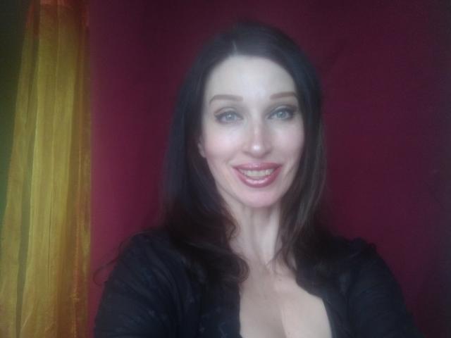 JillianPattie has pale skin, blue eyes, and long brown hair. She's smiling with red lipstick, wearing a black top that shows her cleavage. Her tits are slightly visible.