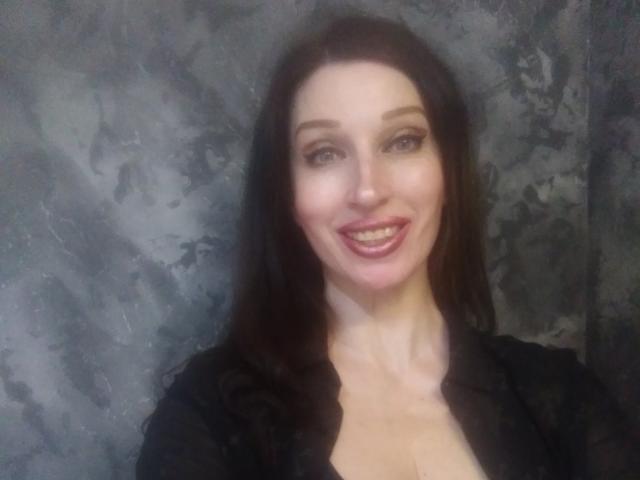 JillianPattie with dark brown hair, blue eyes, and a smile, wearing a black top, showing her tits, against a textured gray background.