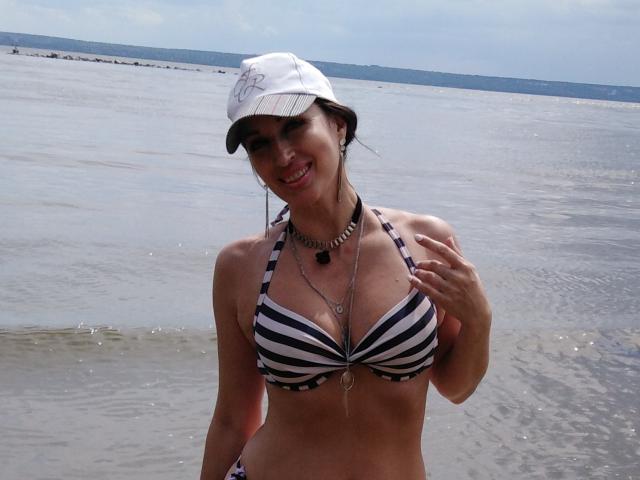 JillianPattie with striped black-and-white bikini, white cap, necklace, smiling, hand on chest, showing her tits, standing in shallow water, looking cute, sunny day, water reflecting on skin.