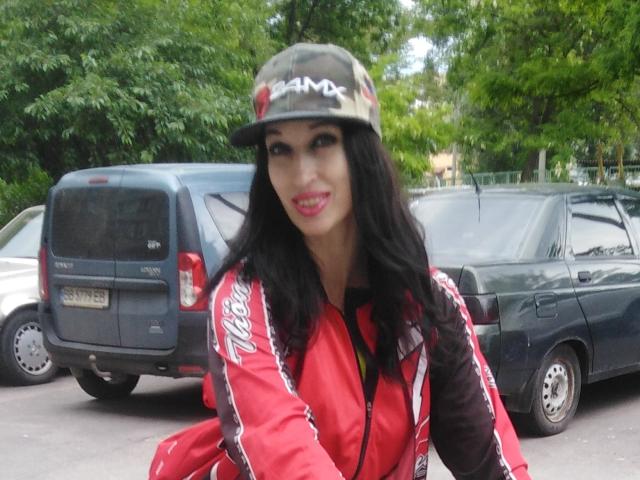 JillianPattie with long black hair, wearing a camouflage cap and red jacket with white stripes, smiling with pink lipstick, standing in front of two dark SUVs.