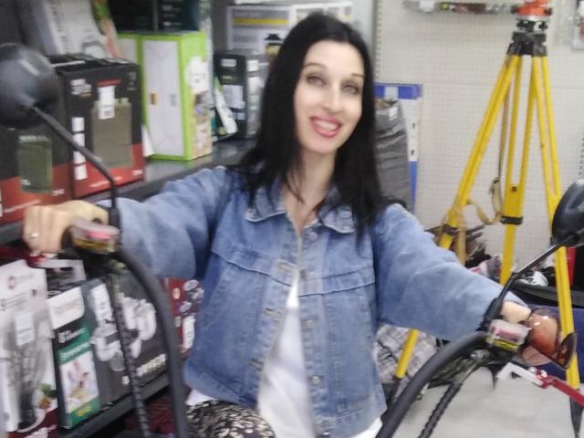 JillianPattie with black hair, blue denim jacket, white shirt, holding bike handlebars, smiling, right arm raised, standing in a store, yellow tripod in background.