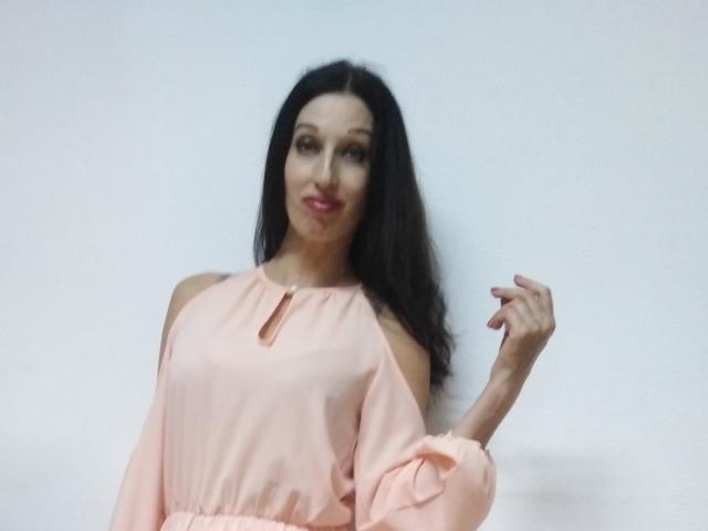JillianPattie with long black hair, pink off-shoulder top, cute smile, and right hand raised, showing off slim fingers. Her pink top has a small keyhole cutout.