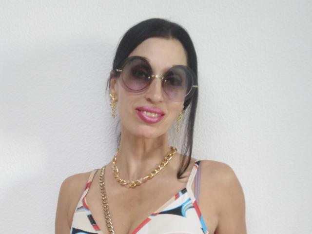 JillianPattie with dark hair, pink lipstick, gold chain necklace, gold hoop earrings, and large round sunglasses, wearing a colorful patterned top, smiling.