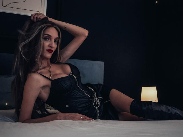 MaiaMoxie with long blonde hair, red lipstick, black leather dress, and boots, laying on a bed, hand in hair, sexy, cute, small breasts, white bed, dim lighting.