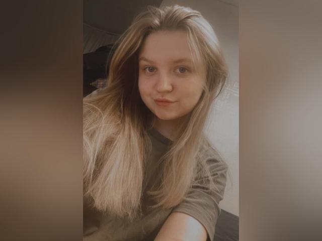 AnitaVine with long blonde hair, soft smile, and blue eyes, wearing a grey shirt, selfie-style, showing subtle makeup and natural beauty.