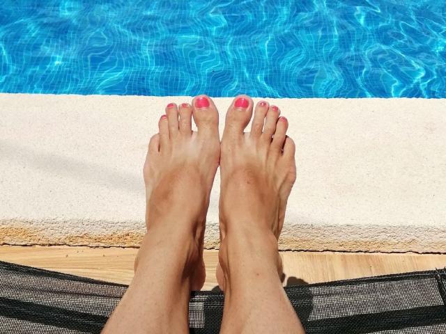 LaFrancaise's feet with pink toenail polish, toes slightly curled, resting on a black mesh lounge chair, next to a bright blue, rippling swimming pool.